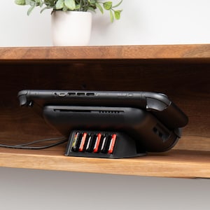 May include: Black gaming console charging dock with red and white game cartridge slots. The console is resting on a black base, with a black cable extending from the side. A small potted plant sits on a wooden shelf above.
