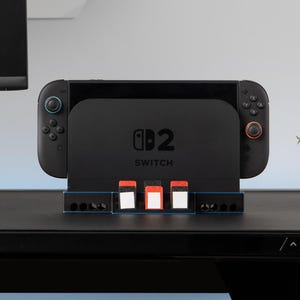Może przedstawiać: Czarna stacja dokująca Nintendo Switch z logo Switch i napisem "Switch" poniżej. Stacja dokująca ma gniazda na kartridże do gier. Kontrolery są czarne z niebieskimi i czerwonymi akcentami.