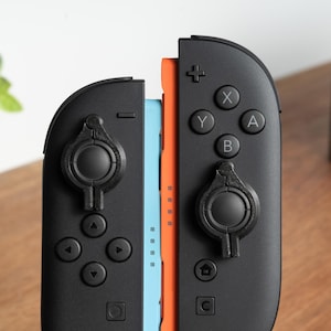 May include: Close-up of two black game controllers with coloured buttons and joysticks. The controllers are connected to a blue and orange central component. The controllers have a variety of buttons, including X, Y, A, and B, and directional pads.