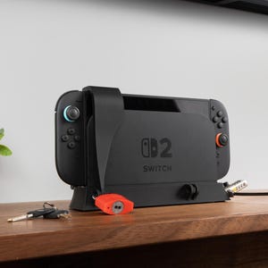 May include: Black Nintendo Switch console with a security lock and keys. The console is secured in a black docking station with the text "Switch". An orange lock is attached to the docking station, with two keys lying on the wooden surface.