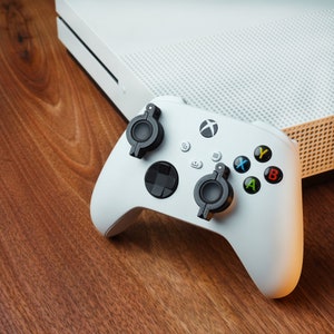 Peut inclure: Une manette blanche Xbox Series X avec des boutons noirs et deux attaches de pagaie noires. La manette est sur une surface en bois.