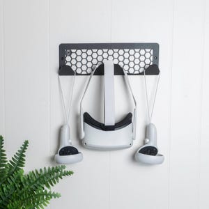 May include: A black metal wall mount with a honeycomb pattern holds a white virtual reality headset and two white controllers.