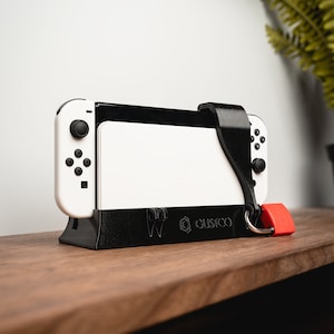 Puede incluir: Una consola Nintendo Switch negra y blanca con un soporte negro y un candado rojo sujeto a la correa. La consola está sobre una superficie de madera.