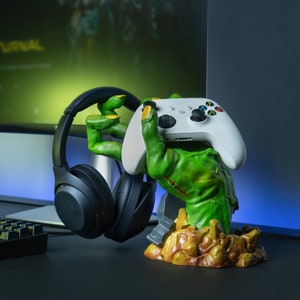 May include: A green zombie hand-shaped gaming accessory holds a white game controller and black headphones. The accessory has yellow nails and a stitched wound. The base is brown and yellow, resembling a rocky terrain. A gaming setup is in the background.