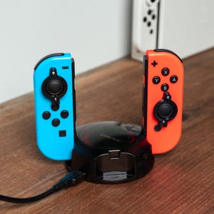 Op de afbeelding: Een zwarte laaddock met twee controllers, een blauwe en een rode, aangesloten. De dock heeft een ronde basis met een zwart-wit ontwerp.