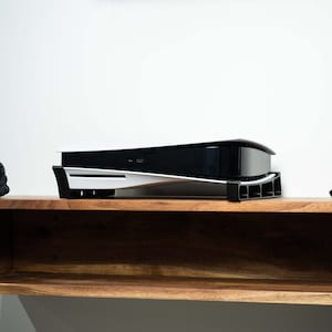 May include: A white and black video game console with a curved design sits on a wooden shelf. The console is resting on a black stand with a white trim.