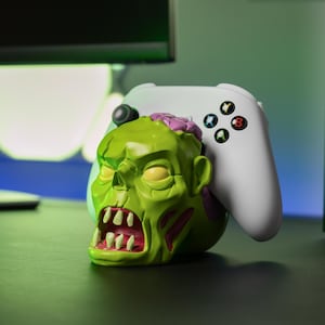 Può includere: Un supporto per controller a forma di testa di zombie verde per un controller Xbox bianco. Lo zombie ha un cervello rosa e una bocca spalancata con denti affilati.
