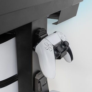 Op de afbeelding: Een witte en zwarte gamecontroller is gemonteerd op een zwarte standaard. De controller heeft een strak ontwerp met meerdere knoppen en joysticks. De standaard lijkt deel uit te maken van een grotere game-opstelling, met een console zichtbaar op de achtergrond.