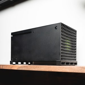 Könnte beinhalten: Eine schwarze Spielekonsole mit einem einzigartigen wabenförmigen Design an der Seite. Die Konsole steht auf einem Holzregal.