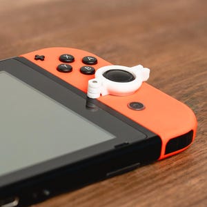 May include: A close-up of a bright orange Nintendo Switch console with a white joystick attachment. The console has black buttons labelled "X", "Y", "A", and "B".