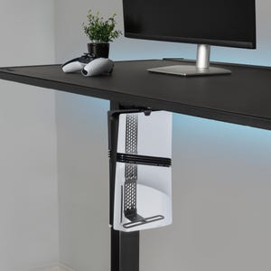 May include: A black desk with a mounted white gaming console holder. A black monitor and a small potted plant sit on the desk. A white and black game controller is also on the desk.
