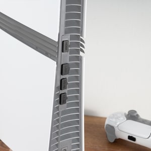 Op de afbeelding: Een witte gameconsole met een zilveren ventilatieopening aan de zijkant. De console heeft vier poorten met de labels HDMI, LAN, USB-A en USB-C. Een witte gamecontroller bevindt zich op de voorgrond.