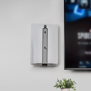 May include: A white gaming console mounted to a wall using a black metal bracket. The bracket features a distinct honeycomb pattern and runs vertically along the centre of the console, securing it firmly in place next to a television screen.