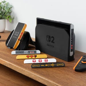 May include: A black Nintendo Switch dock and controller with several decorative controller skins. The skins feature pixelated hearts, mushrooms, and other gaming-themed designs. The Switch dock has the text "Switch" on it.