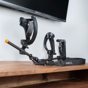 May include: A black VR headset and controllers are mounted on a wall-mounted stand. A black and orange VR accessory is attached to a black strap. The strap is labelled "GUSTCO".