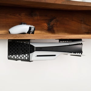 Könnte beinhalten: Ein schwarzes Metallregal mit einer weißen Spielekonsole, die darunter montiert ist. Die Konsole ist mit einer schwarzen Halterung am Regal befestigt. Ein weißer Videospielcontroller liegt auf dem Regal.