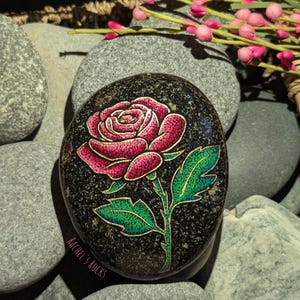 May include: A smooth, oval-shaped black stone featuring a hand-painted rose with vibrant pink petals and green leaves, outlined in gold. The surface has a speckled texture, and the text RACHEL'S ROCKS is printed in small letters on the lower left side.