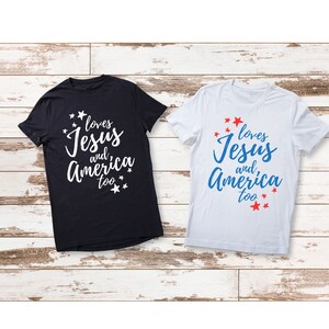 Könnte beinhalten: Zwei T-Shirts mit dem Aufdruck "loves Jesus and America too". Ein T-Shirt ist schwarz mit weißem Text und das andere ist weiß mit blauem und rotem Text.