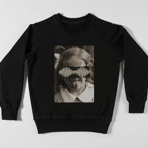 Może przedstawiać: Czarna bluza z nadrukiem graficznym przedstawiającym dziewczynę z falą dźwiękową nad oczami. Fala dźwiękowa to logo zespołu "The 1975".