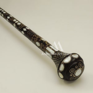 May include: A dark brown walking stick with a decorative, rounded knob. The knob is inlaid with white mother-of-pearl and gold accents. The stick is decorated with a geometric pattern of white mother-of-pearl and gold.