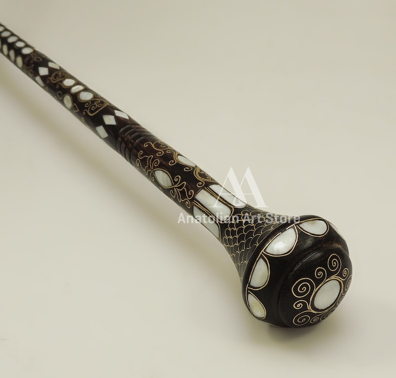 May include: A dark brown walking stick with a decorative, rounded knob. The knob is inlaid with white mother-of-pearl and gold accents. The stick is decorated with a geometric pattern of white mother-of-pearl and gold.