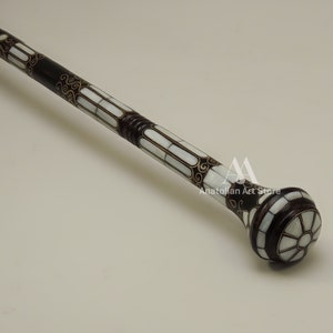 May include: A decorative walking stick with a dark brown wooden shaft and a white mother-of-pearl inlay. The shaft is decorated with intricate gold floral patterns. The top of the stick is a round knob with a white mother-of-pearl inlay and a gold floral pattern.