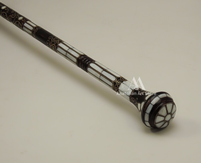May include: A decorative walking stick with a dark brown wooden shaft and a white mother-of-pearl inlay. The shaft is decorated with intricate gold floral patterns. The top of the stick is a round knob with a white mother-of-pearl inlay and a gold floral pattern.