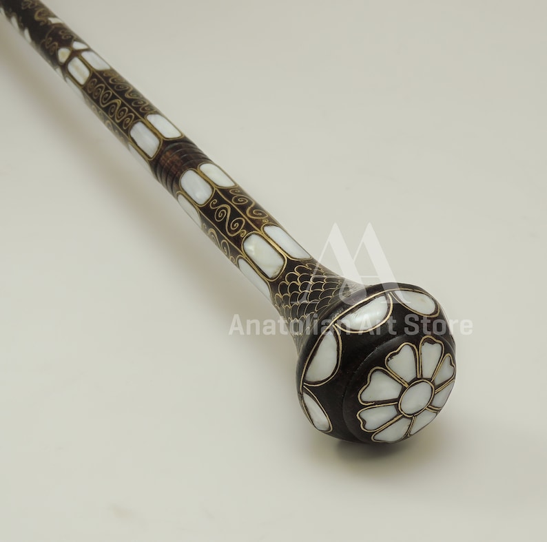 May include: A decorative walking stick with a dark brown wooden shaft and a white mother-of-pearl inlay design. The top of the stick is a round knob with a white flower design.