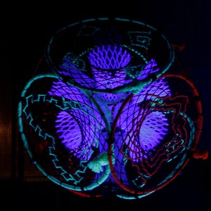 May include: Three dreamcatchers with intricate woven patterns are illuminated in a dark room with a purple glow. The dreamcatchers are made of different colored threads, including red, blue, and green.