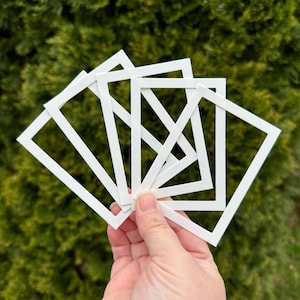 May include: A hand holding several white picture frame mats against a backdrop of green foliage. The mats are rectangular with a square opening in the center, designed for displaying photographs or artwork. The mats are clean and crisp.