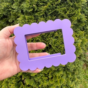 May include: A light purple, scalloped-edge picture frame. The frame is rectangular with a cutout center. The frame is held in front of a green bush.