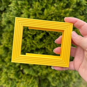 May include: A bright yellow rectangular picture frame with a layered design. The frame is held by a hand against a blurred green background. The frame's edges have a series of parallel lines, creating a textured appearance.