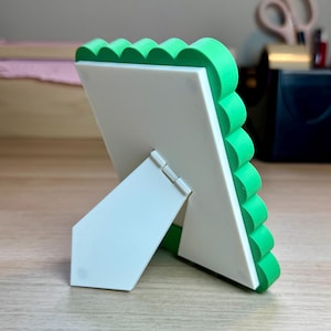 May include: A white picture frame with a green scalloped edge, propped up on a white stand. The frame is made of a smooth material and is set against a light wooden surface. The back of the frame is visible.