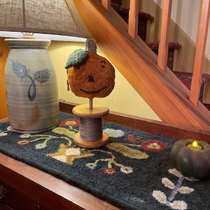 May include: A hand-stitched felt pumpkin with a smiling face, a green felt stem, and a wooden spool base. The pumpkin is displayed on a black and white rug with floral designs.