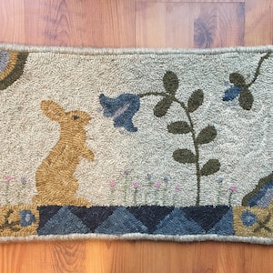 May include: A beige rug with a brown rabbit, a blue flower, and green leaves. The rug has a blue and gray border.