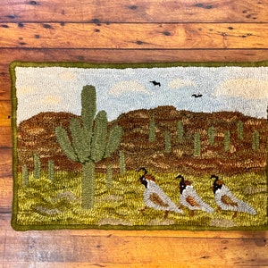 Op de afbeelding: Een handgestikt tapijt dat een woestijnscène voorstelt met een grote cactus, kleinere cactussen en drie kwartels op de voorgrond. Het tapijt heeft een groene rand en een lichtblauwe lucht.