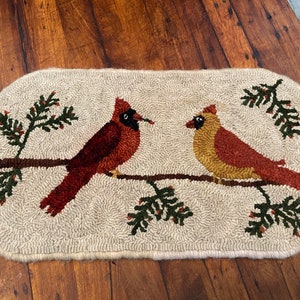 May include: An oval-shaped rug featuring two cardinal birds perched on a brown branch with green pine needles and red berries. The birds are red and tan, set against a beige background. The rug is made of a textured, looped material.