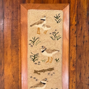 May include: A framed needlepoint rug with a beige background featuring three brown and white birds with black eyes. The birds are surrounded by green grass and brown branches. The rug is framed in a dark wood frame.