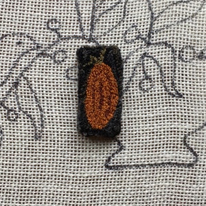 May include: A small, brown, felt pumpkin with a green stem, stitched on a white canvas.