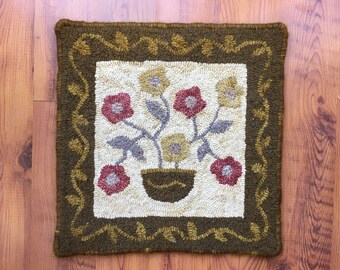Könnte beinhalten: Ein handgestickter Teppich mit einem Blumendesign. Der Teppich zeigt einen braunen Topf mit fünf Blumen in Rot-, Gelb- und Grüntönen. Der Teppich ist von einem braunen Rand mit einem Rankenmuster umgeben.