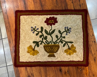 Könnte beinhalten: Ein handgestickter Teppich mit einem Blumendesign. Der Teppich zeigt eine braune Vase mit gelben, roten und blauen Blumen. Der Teppich hat einen roten und braunen Rand.