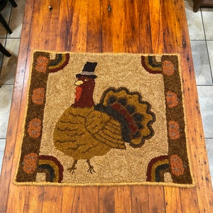May include: A brown and beige hooked rug with a turkey wearing a top hat in the center. The turkey is surrounded by a brown border with orange and brown accents.