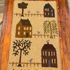 Op de afbeelding: Een handgestikt tapijt met een grillig ontwerp met drie huizen, bomen en vogels. De huizen zijn bruin, groen en blauw met witte ramen. De bomen zijn groen en bruin. De vogels zijn blauw. Het tapijt heeft een beige achtergrond en een bruine rand.