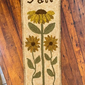 Könnte beinhalten: Ein beiger Teppich mit braunem Rand zeigt drei Sonnenblumen mit grünen Stielen und Blättern. Das Wort "Herbst" ist in Braun in der Mitte oben auf dem Teppich gestickt.