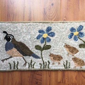 May include: Hand-hooked rug featuring a quail with chicks and blue flowers. The rug has a neutral background with a border. The quail is brown, blue, and tan, and the flowers have yellow centers. The rug is rectangular.