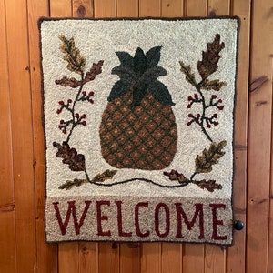 Könnte beinhalten: Eine cremefarbene Willkommensmatte mit einem braunen Ananas-Design und dem Wort "WELCOME" in roten Buchstaben.