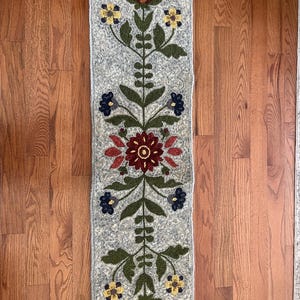 May include: A rectangular, grey wool rug with a floral design. The rug features a central stem with symmetrical branches, adorned with burgundy, yellow, and blue flowers. The rug's design is reminiscent of folk art.