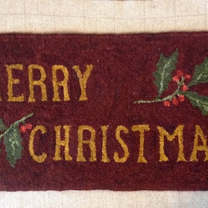 May include: A maroon-coloured rug with the words "Merry Christmas" embroidered in gold thread. The rug features two sprigs of holly with red berries.