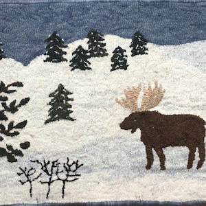 May include: A brown moose with large antlers stands in a snowy landscape. The scene is depicted in a needlepoint rug with a blue sky and green trees.