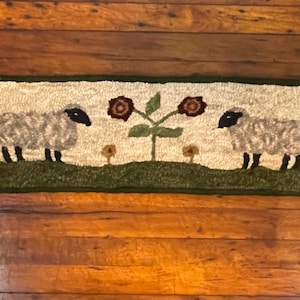 May include: A white and green rug featuring two sheep and a plant with two flowers. The sheep are facing each other and the plant is in the centre of the rug.